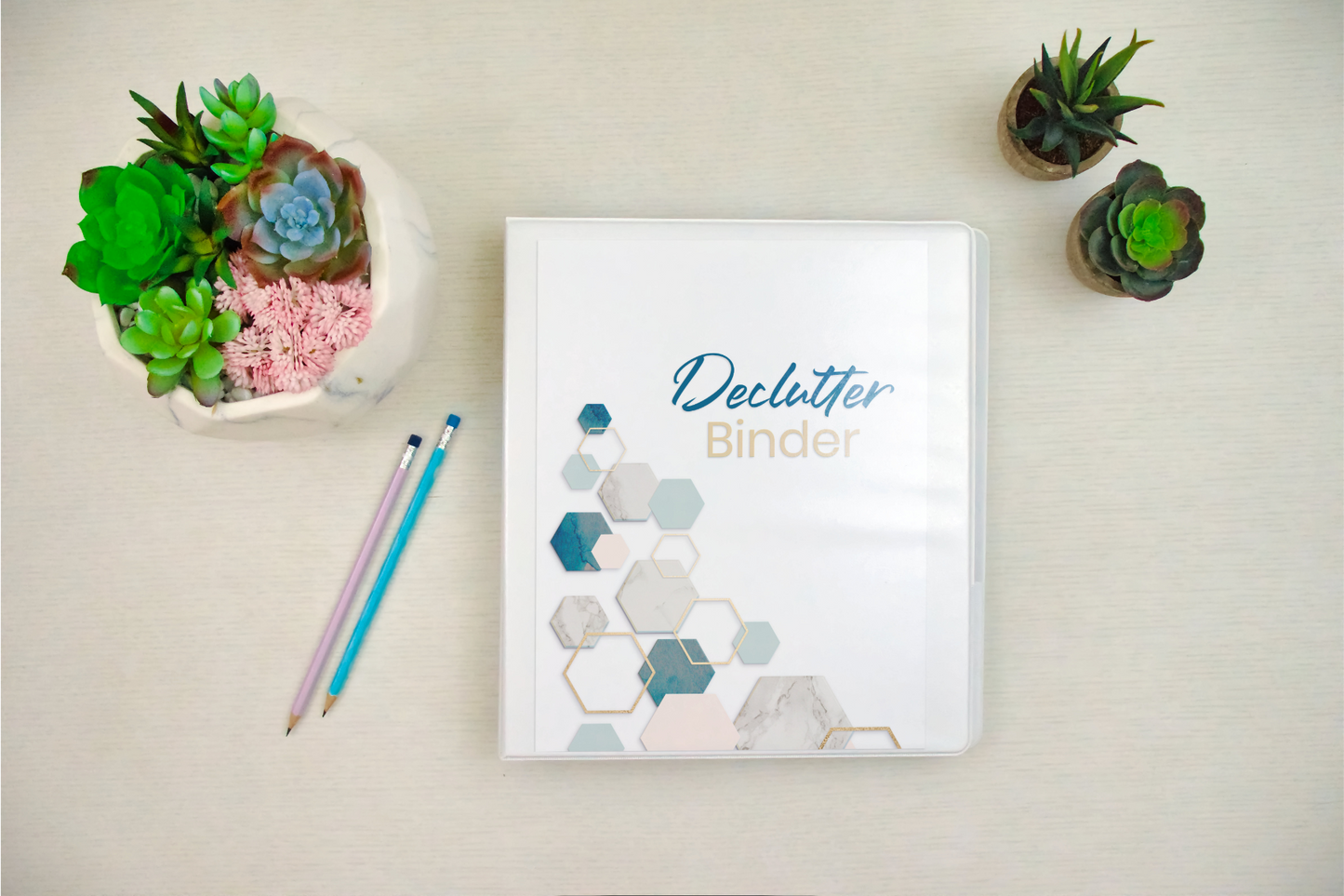 The Wondermom Shop's "Declutter Binder: Your Guide to a Tidy and Stress-Free Home" with hexagon designs rests on a desk with two pencils and small succulent plants, featuring room-by-room checklists for easy organization.