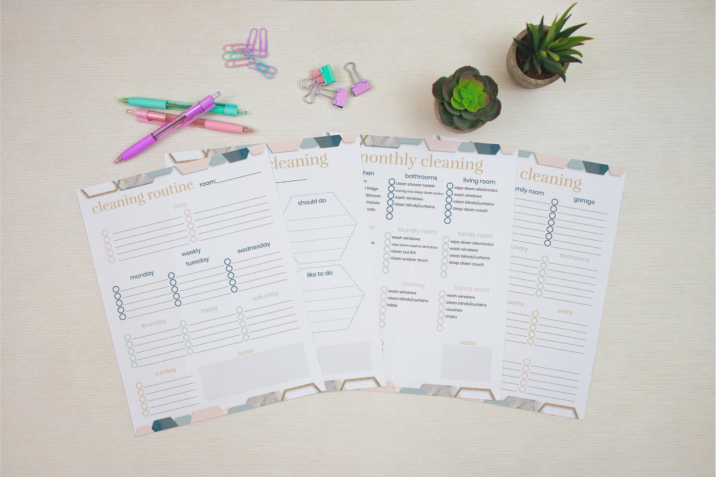 Four copies of the "Cleaning Binder: Your Go-To Guide for a Spotless Home" by Wondermom Shop are laid out on a table, accompanied by colorful pens, paper clips, and a small plant in the background.