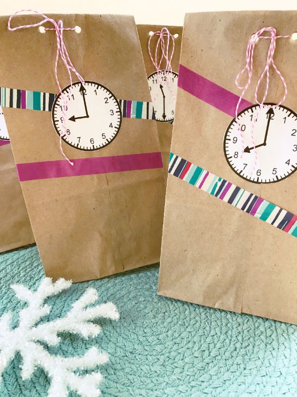VIP Vault's New Year Countdown Activity for Kids features brown paper bags with clock face tags, pink string, and striped tape. A white snowflake ornament on a teal woven surface adds magical fun to the festive occasion.