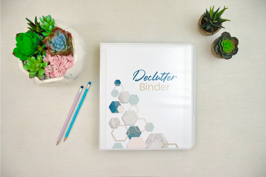 The Wondermom Shop's "Declutter Binder: Your Guide to a Tidy and Stress-Free Home" with hexagon designs rests on a desk with two pencils and small succulent plants, featuring room-by-room checklists for easy organization.