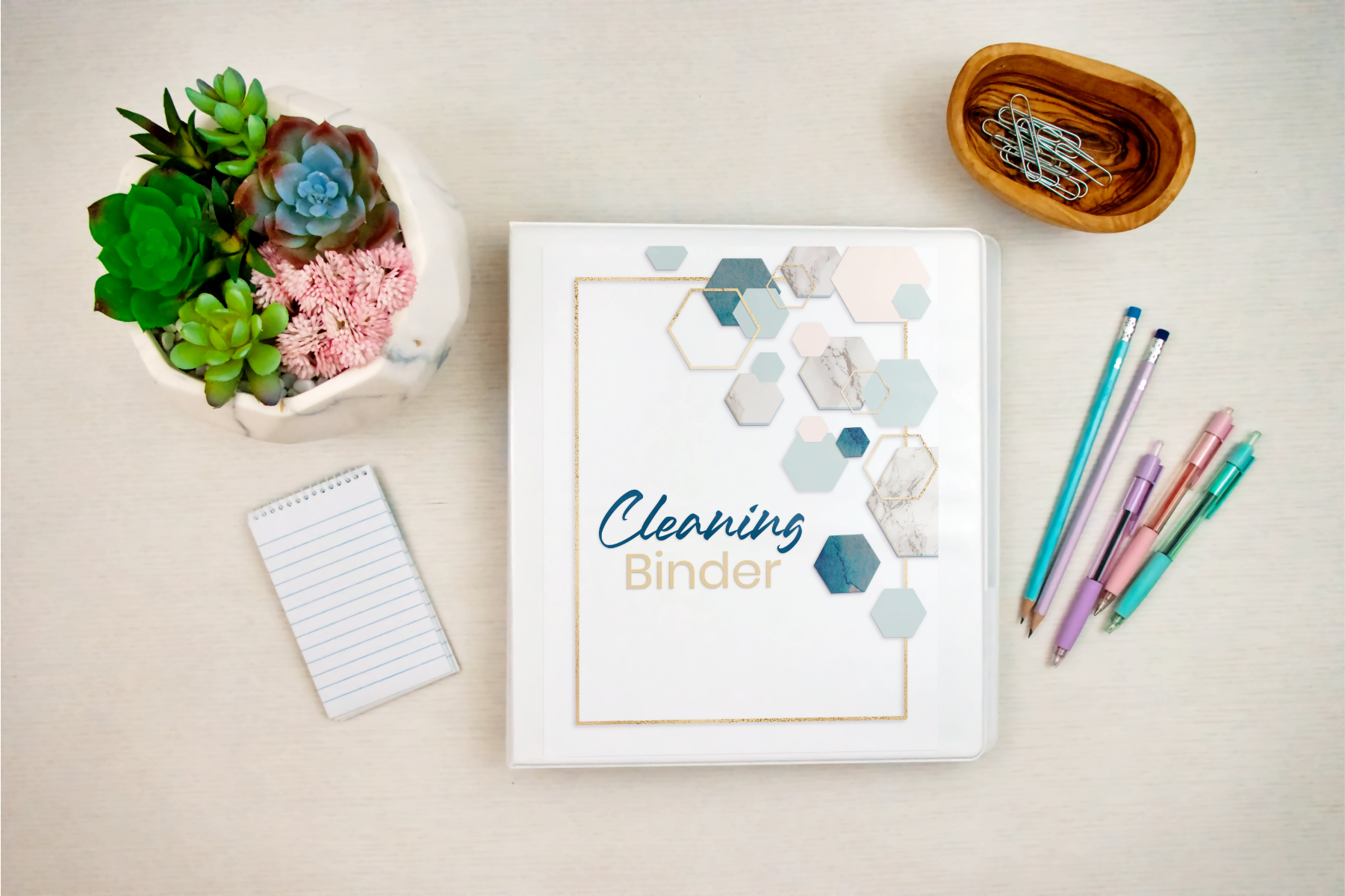 The "Cleaning Binder: Your Go-To Guide for a Spotless Home" by Wondermom Shop rests on a desk beside a succulent bowl, small notepad, colored pens, and a wooden bowl of paperclips.