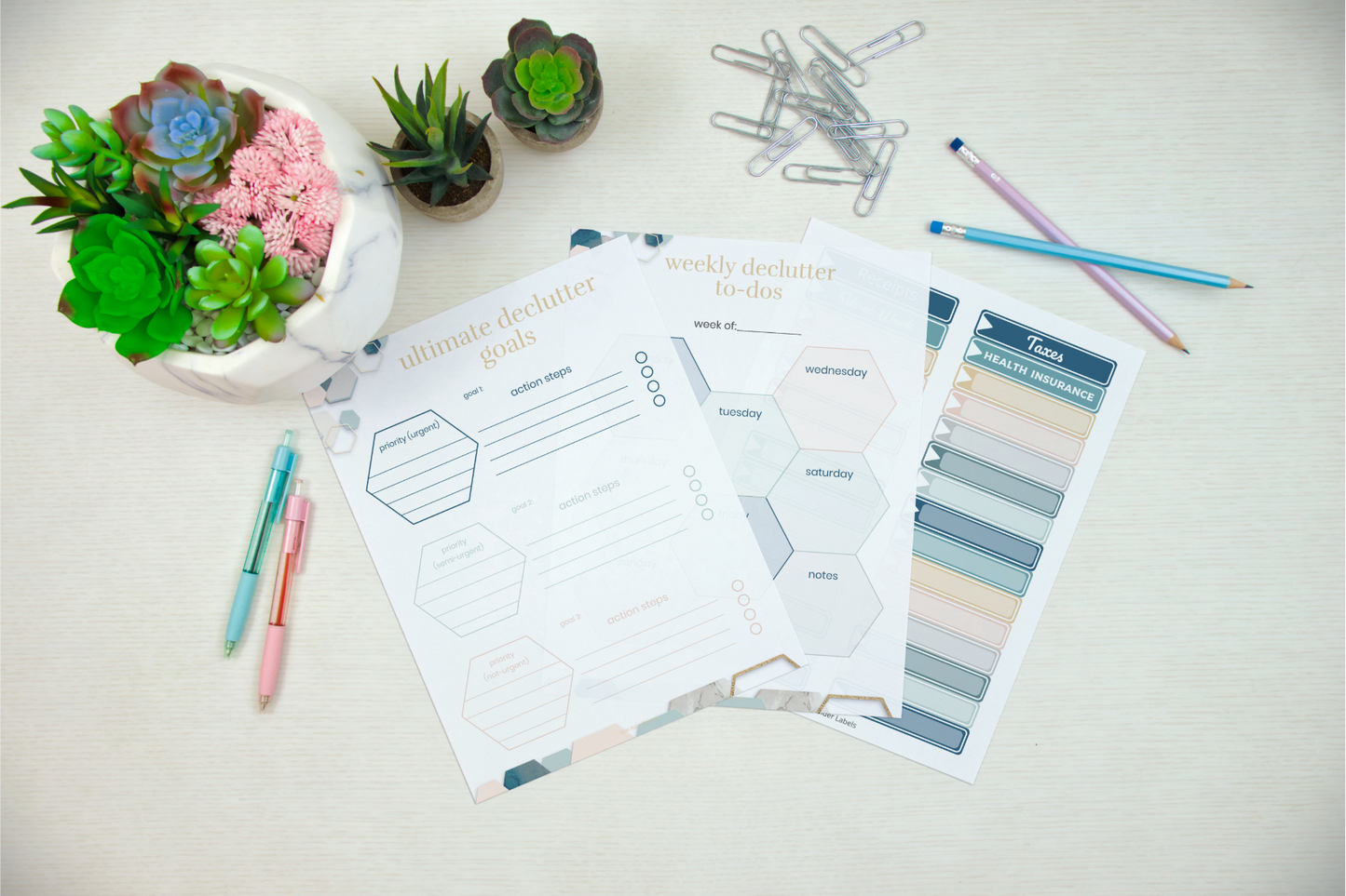Overhead view of a desk with colorful pens, paper clips, succulent plants, and planner pages marked "ultimate declutter goals" alongside your Wondermom Shop Declutter Binder: Your Guide to a Tidy and Stress-Free Home—ideal for simplifying your space with room-by-room checklists and weekly tasks.