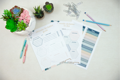 Overhead view of a desk with colorful pens, paper clips, succulent plants, and planner pages marked "ultimate declutter goals" alongside your Wondermom Shop Declutter Binder: Your Guide to a Tidy and Stress-Free Home—ideal for simplifying your space with room-by-room checklists and weekly tasks.