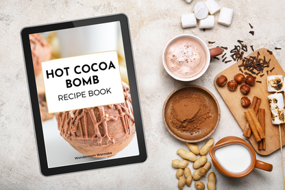 A tablet featuring the "Hot Cocoa Bombs Recipe Book" by Wondermom Wannabe sits beside a cup of hot chocolate, cocoa powder, milk, cinnamon sticks, marshmallows, and nuts on a tabletop.