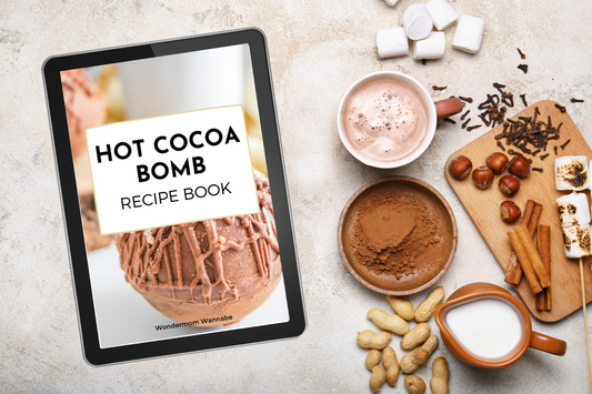 A tablet featuring the "Hot Cocoa Bombs Recipe Book" by Wondermom Wannabe sits beside a cup of hot chocolate, cocoa powder, milk, cinnamon sticks, marshmallows, and nuts on a tabletop.