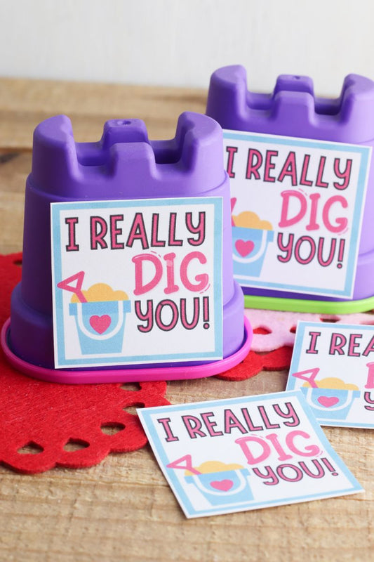 Two purple sandcastle molds, ideal for a VIP Vault Kinetic Sand Valentine, include "I Really Dig You" cards. Arranged on a red felt mat with extra Valentine's Day printables, it's an enchanting gift idea.